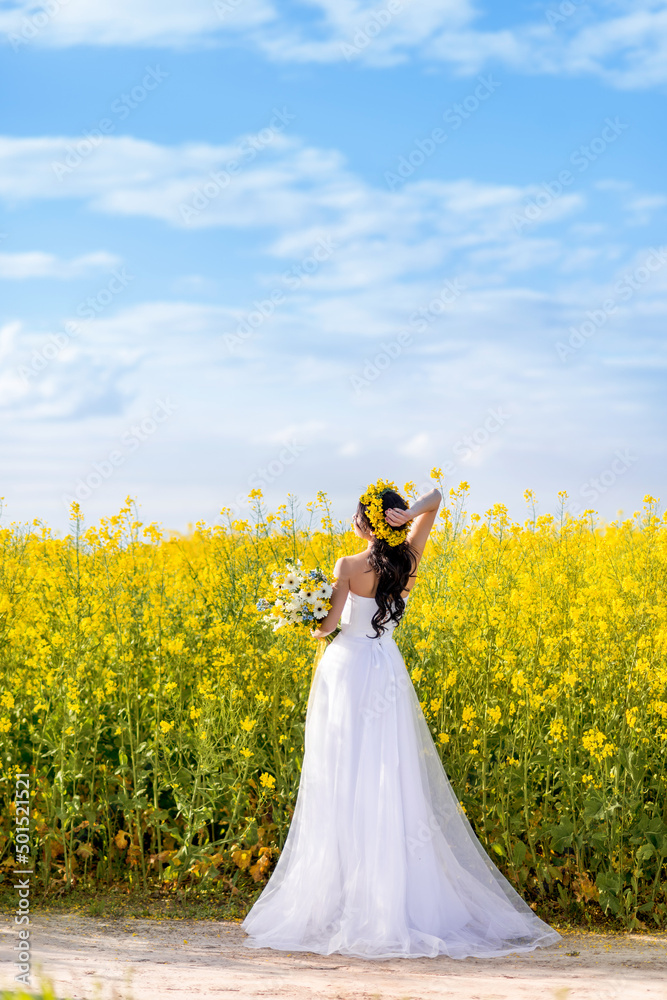 young beautiful woman in a long white dress is standing on a rural country road. A bride with a bouquet in a yellow flowering rapeseed field. Against a beautiful blue sky