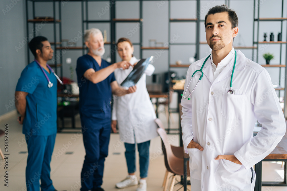 Portrait of confident young male doctor in white uniform standing in medical meeting office, looking at camera. Multiethnic team of physician working in background, discussing MRI scan of patient. Portrait of confident young male doctor in white uniform standing in medical meeting office, looking at camera. Multiethnic team of physician working in background, discussing MRI scan of patient.
