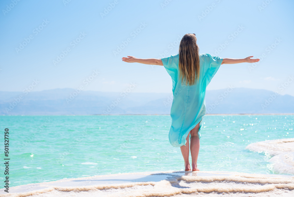 Young woman in turquoise flying pelerine stands by back on white salt island Dead sea and gets enjoy. Space for text. Travel and vacation in summer in Israel Young woman in turquoise flying pelerine stands by back on white salt island Dead sea and gets enjoy. Space for text. Travel and vacation in summer in Israel