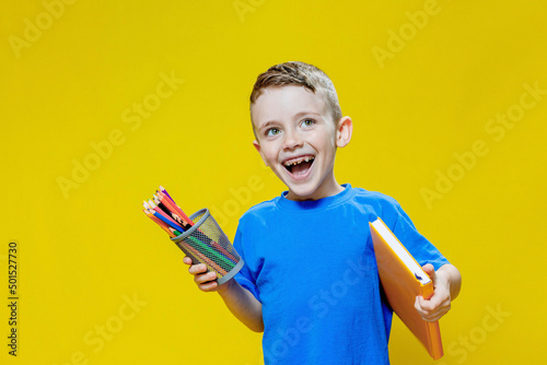 Smiling happy schooler in blue t-shirt holding multicolored pencils and book on yellow background