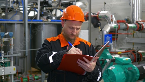 An industrial operator of the oil and gas complex, a man with a beard in an orange helmet, checks equipment with records of readings in a log, in a workshop with power plants for pumps and compressors © ShantiMedia