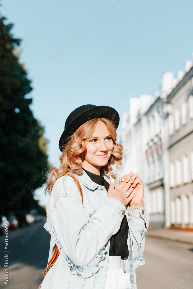 Fototapeta premium Pretty young hipster woman in hat standing on street in city and enjoying walk. Woman model with curly hair and cute smile posing on sunny day outdoors. Vertical view