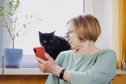 Senior blonde woman using mobile phone for video call with friend or children, showing black pet cat, sitting near window at home. Online consultation with the veterinarian using wireless 5g internet