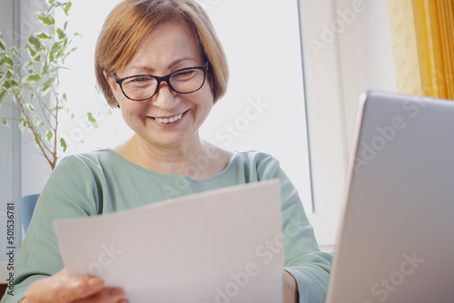 Happy middle aged elderly woman in eyeglasses reading paper document or postal letter from friend, sitting with laptop at home. Rejection of pleasant paperwork bank notice or post correspondence