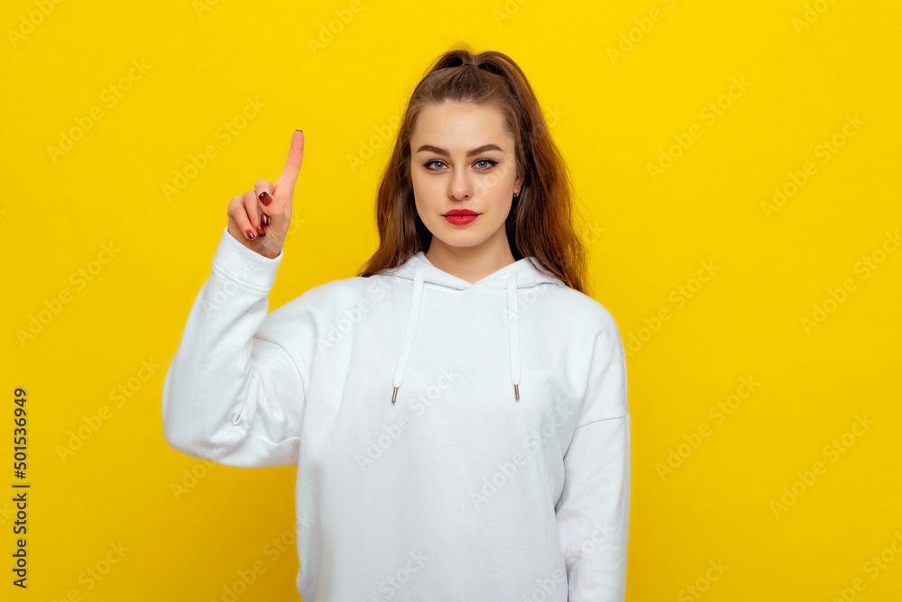 Serious beautiful brunette girl in white casual style sweatshirt, frowning, pointing fingers up, standing over yellow background Serious beautiful brunette girl in white casual style sweatshirt, frowning, pointing fingers up, standing over yellow background