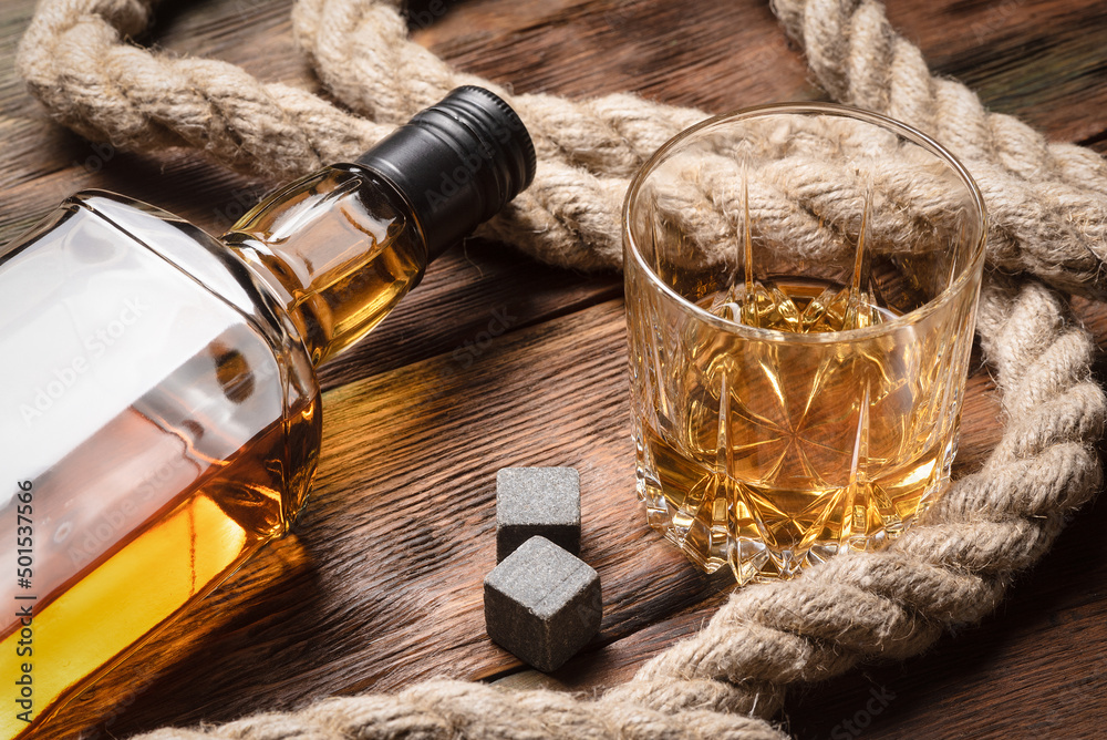 Fototapeta premium Whiskey in the drinking glass, bottle and mooring rope on the brown wooden table background.