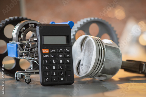 Car repair costs concept. Automobile spare parts cost estimate. Calculator, engine piston and bearings in the shopping cart on the metal workbench background close up.