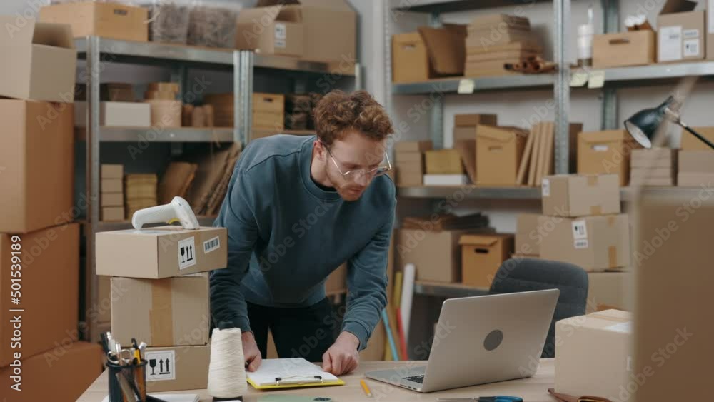 Ginger caucasian man standing at the table, looking at the laptop screen and writing information about parcel delivery while working at the warehouse. Post service and small business concept. Ginger caucasian man standing at the table, looking at the laptop screen and writing information about parcel delivery while working at the warehouse. Post service and small business concept.