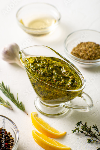 Chimichurri sauce in a gravy bowl on a white background. Various spices lie nearby. Argentinean vegetarian sauce made from olive oil, oregano, parsley.