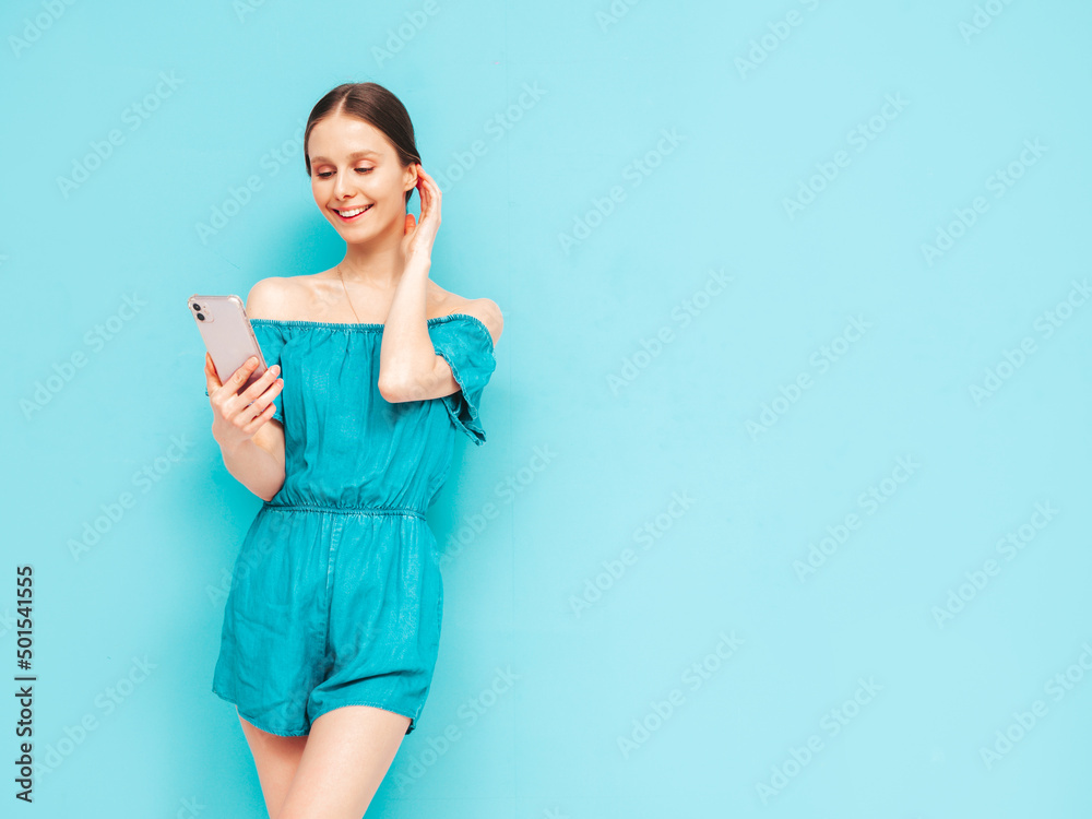 Young beautiful smiling female in trendy summer blue overalls. Sexy carefree woman posing near wall in studio. Positive brunette model looking at cellphone screen. Holding smartphone and using apps Young beautiful smiling female in trendy summer blue overalls. Sexy carefree woman posing near wall in studio. Positive brunette model looking at cellphone screen. Holding smartphone and using apps