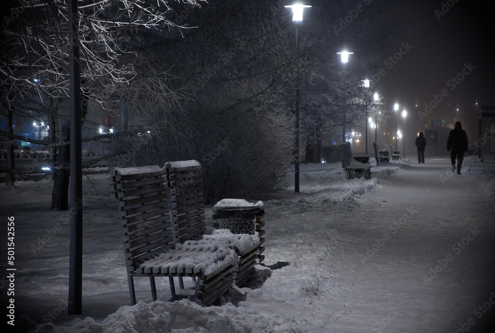 Winter night avenue benches covered in snow with two people silhouettes walking away