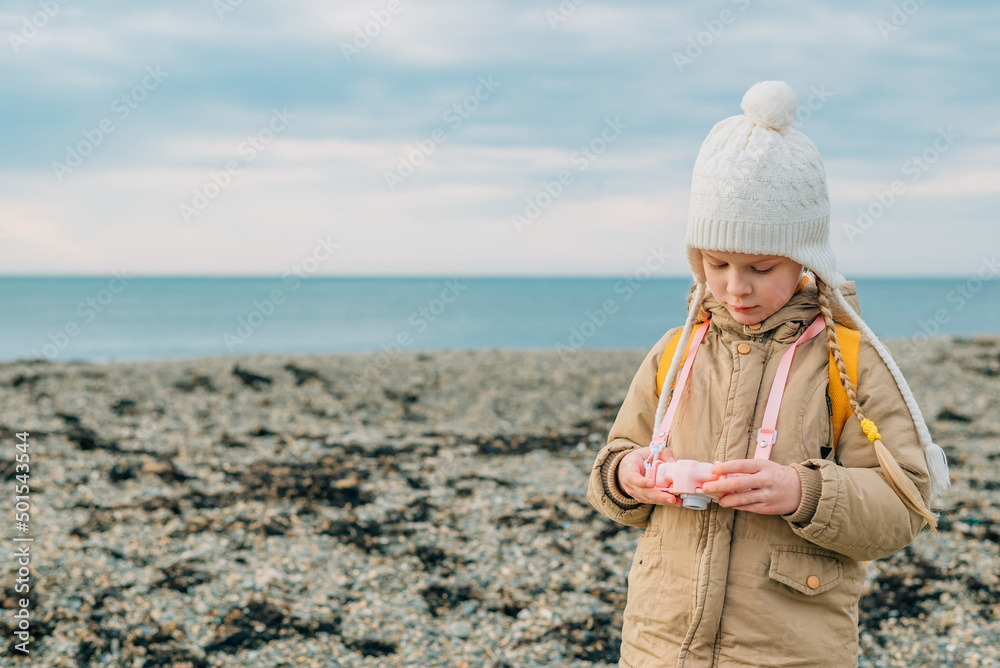little girl looks at the screen of a children's pink camera to see her photos of the autumn, spring seascape. the child is passionate about his hobby: photography