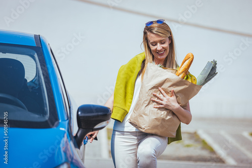 Young woman riding shopping cart full of food on the outdoor parking. Young woman in car park, loading shopping into boot of car. Shopping successfully done. Woman putting bags into car after shopping