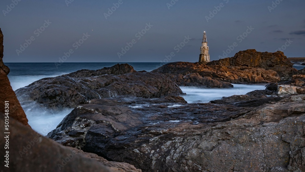 Attractive night seascape, sea waves splashing over rocks, shining lighthouse. Evening darkness, long exposure, blurry water trails and misty steam. Southern Black Sea coast, Ahtopol, Bulgaria.