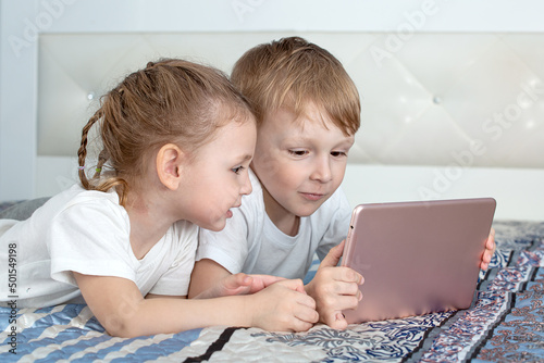 Kids and gadgets. A boy and a girl are lying on the bed, looking at the tablet and smiling
