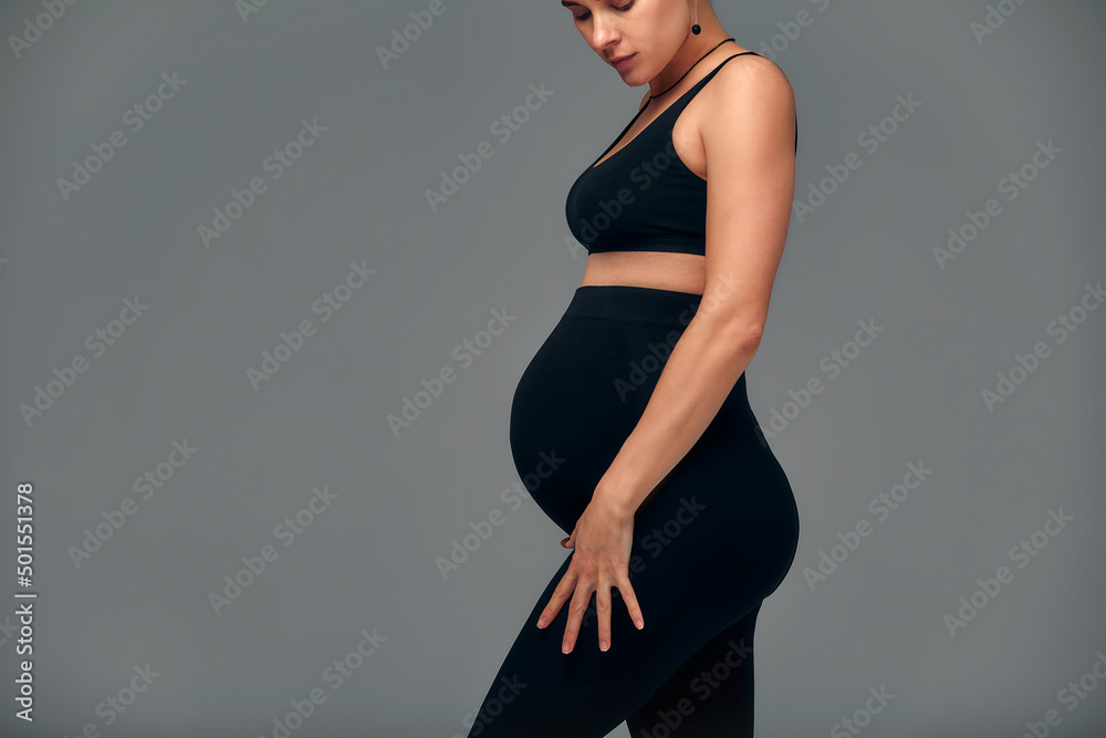Cute pregnant belly isolated on gray. Side view of young pregnant woman embracing her abdomen with hands. Big belly on the third trimester of pregnancy close-up. Concept of pregnant life. Cute pregnant belly isolated on gray. Side view of young pregnant woman embracing her abdomen with hands. Big belly on the third trimester of pregnancy close-up. Concept of pregnant life.