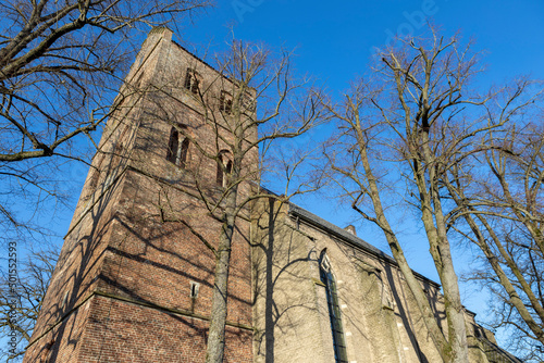 Church tower of Christophorus kerk in Geesteren town seen from below with winter barren tree branches flanking the authentic small village religious building against a clear blue sky photo
