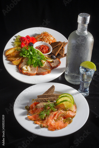 Close-up view of bottle of vodka and glasses with Marinated vegetables and sliced meat decorated with parsley and dill, also appetizer - salted salmon and bread at the plate standing on black