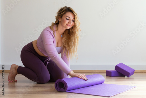Young woman rolling lilac mat after gymnastics, yoga or Pilates sports exercises at home or in the studio. A side view of a slender, positive blonde girl on floor near a window on white background.