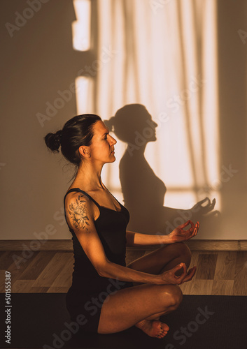 Front view of young sporty woman practicing yoga meditation on black sport mat in at home with sunset or sunrise light with shadows.