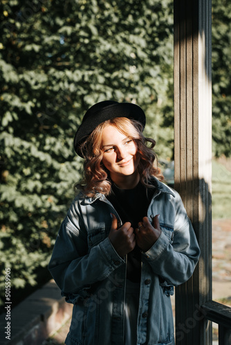 Pretty young woman with curly hair and sweet smile in park on sunny evening. Cute hispter model posing in hat and denim jacket looking away outdoors. Vertical view