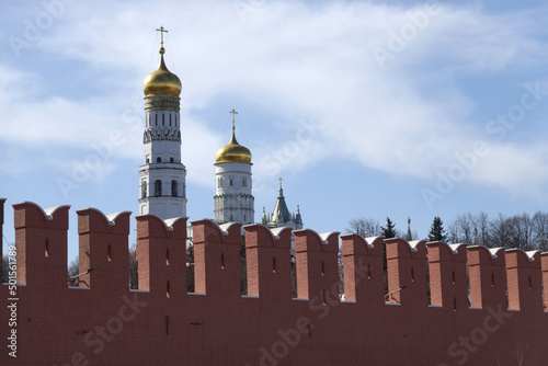 Ivan the Great Bell Tower with golden cupolas behind the red brick Kremlin wall on sunny day