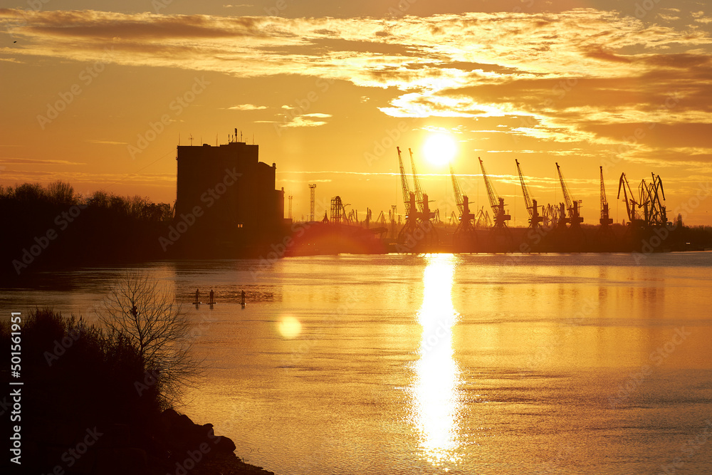 River landscape with silhouette of people paddle on stand up paddle boarding (SUP) and kayak at sunrise on quiet surface of autumn Danube river against port cranes