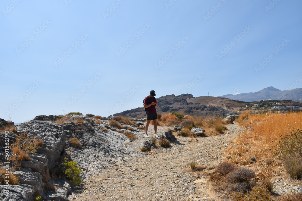 Hiking man standing on mountain top near a trail path and looking at his smartphone