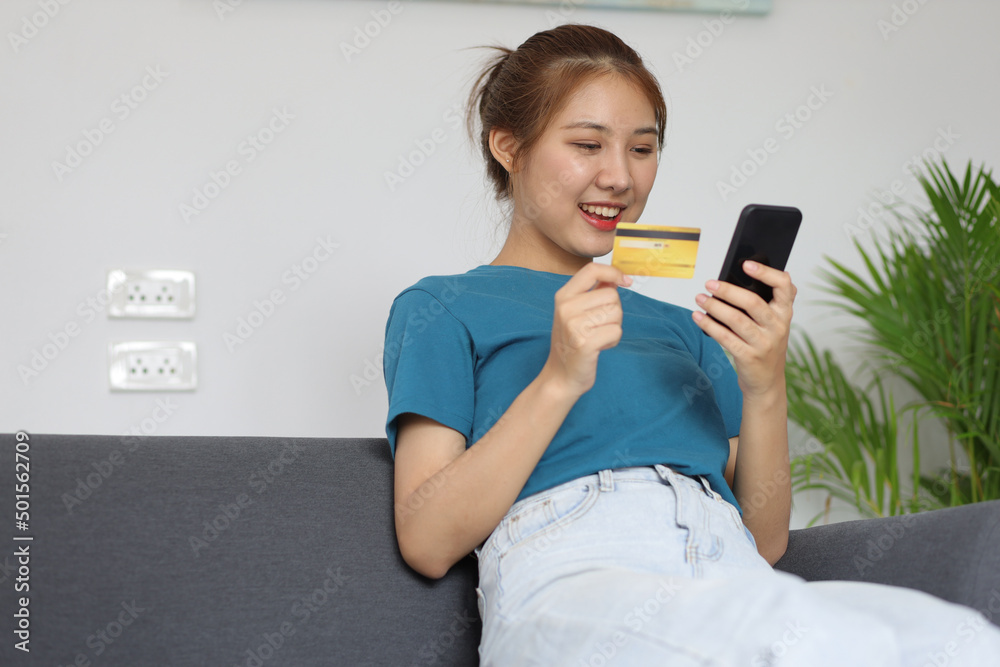Cheerful Asian young woman lifestyle while sitting on sofa at home using mobile phone and having fun. Woman holding credit card and shopping online. Cheerful Asian young woman lifestyle while sitting on sofa at home using mobile phone and having fun. Woman holding credit card and shopping online.