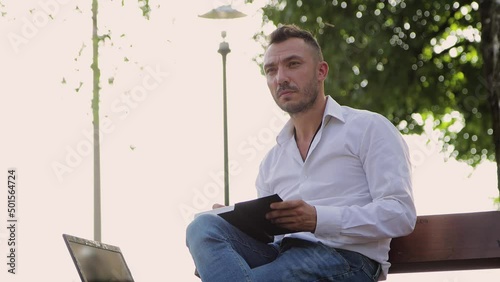 A concentrated man in a white shirt writes down plans in a notebook on a bench. Young man in a city park, hot sunny summer day. Warm soft light, close-up. Against the background of green trees. 4K UHD
