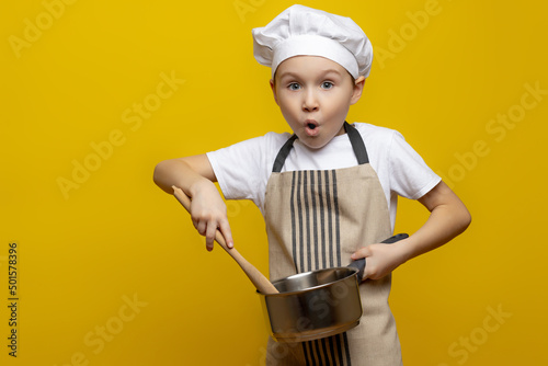 Isolated photo of a surprised European teenage boy in an apron and a chef's hat, holding a saucepan and a wooden spoon in his hands, tasting, cooking a surprise dinner for his parents.