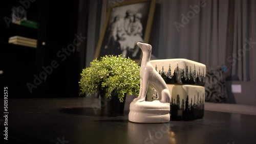 A close-up shot of a miniture white dog sculpture, leafy bonsai tree, and small painted black and cream ceramic box together on a table. A portrait, bookcase, and cusion are visible in the background. photo