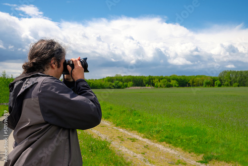Male photographer taking pictures of barley field with forest far away and blue sky with clouds. Early springtime landscape. Germany, countryside in Brandenburg North from Berlin.