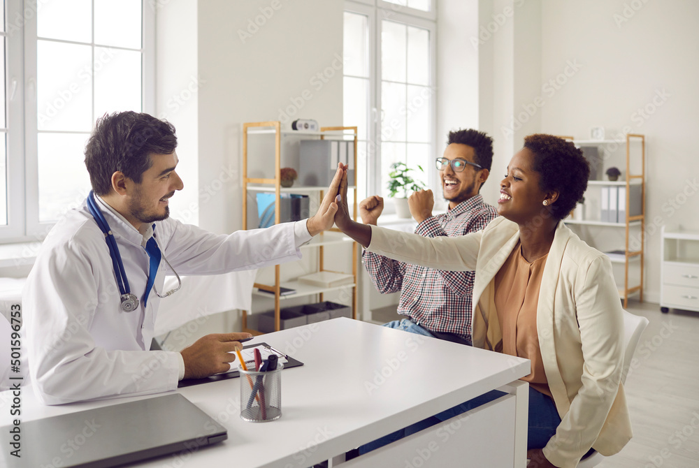 Fototapeta premium Smiling man doctor give high five to happy multiethnic couple at hospital consultation. Happy male GP celebrate successful pregnancy planning or good medical results with excited ethnic young family.