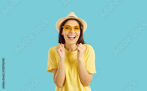 Cheerful beautiful young woman getting excited in anticipation of summer holiday trip. Happy pretty girl in yellow T shirt and hat isolated on blue background looking at camera, smiling and laughing photo