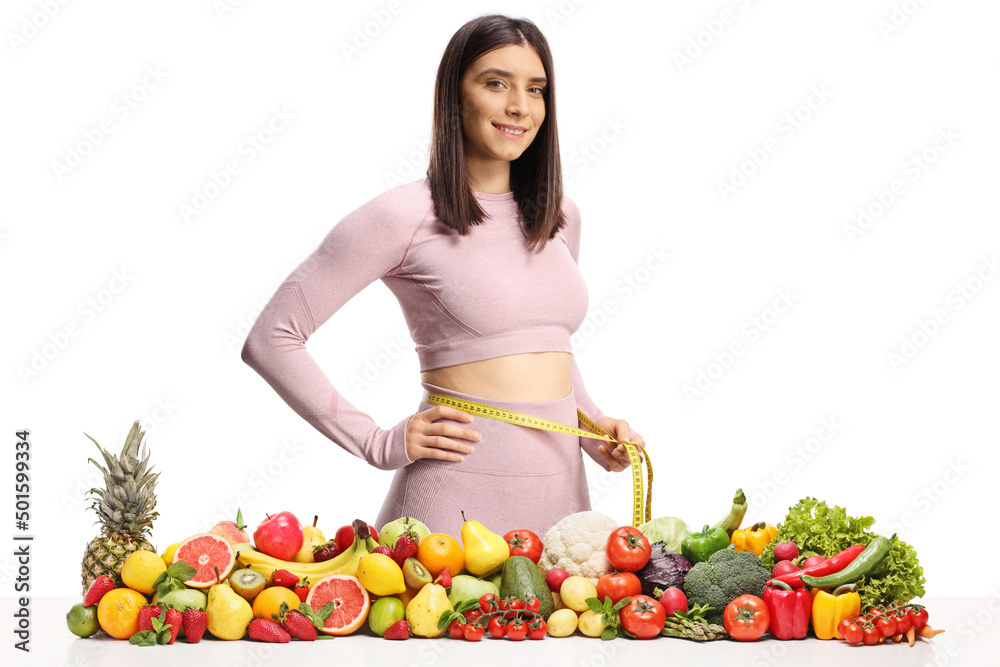 Fit woman in sportswear measuring her waist behind a pile of fruits and vegetables Fit woman in sportswear measuring her waist behind a pile of fruits and vegetables