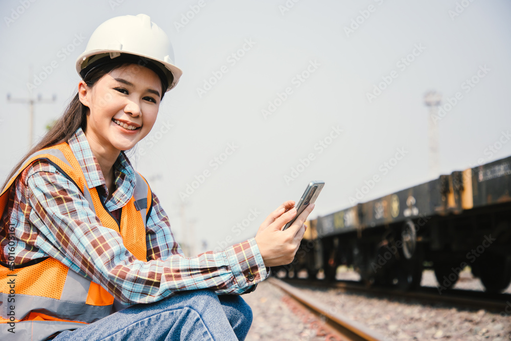 Fototapeta premium Portrait of Asian beautiful engineering using smartphone for control labor worker with wear hardhat in front of train garage. Back view of contractor on background of outdoor old train garage.