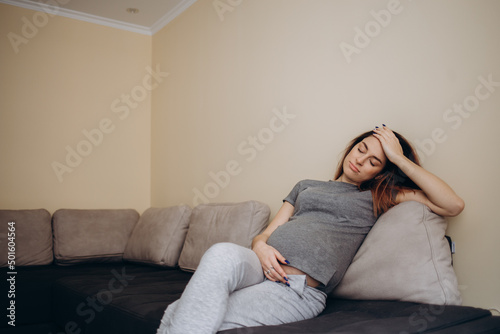 Young beautiful pregnant woman sitting at home on the couch sick, has a headache and nausea