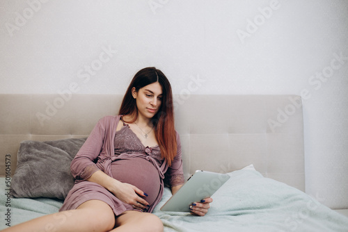 Side view of a young pregnant woman resting with a tablet on the sofa at home. A beautiful pregnant woman who holds her belly and relaxes with a digital tablet in bed during pregnancy.