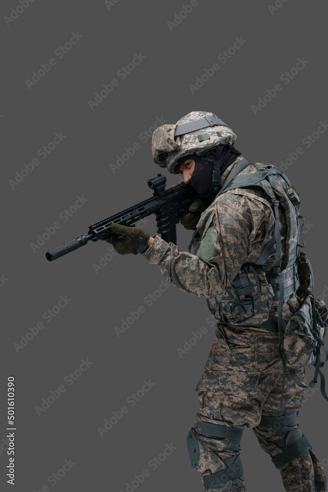 Phot of brave serviceman dressed in vest and uniform aiming rifle against gray background.