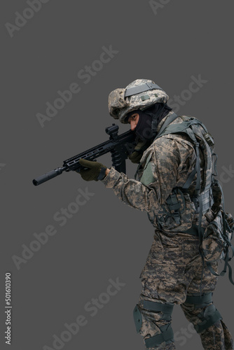 Phot of brave serviceman dressed in vest and uniform aiming rifle against gray background.