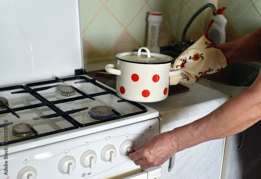 An elderly woman puts a pan on a gas stove with a hand in a mitten