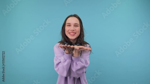 Positive girl in a sweatshirt and headphones around his neck shows a heart gesture with a smile on his face and sends an air kiss and smiles