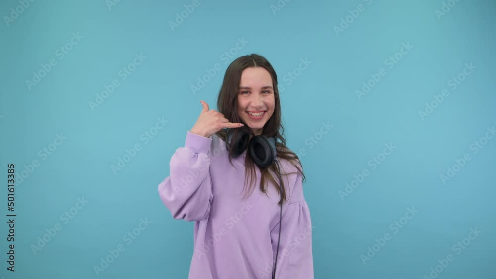 Positive woman in purple clothes and headphones shows a gesture of a call to the camera and and looks with a smile on his face, isolated on a blue background. Positive woman in purple clothes and headphones shows a gesture of a call to the camera and and looks with a smile on his face, isolated on a blue background.