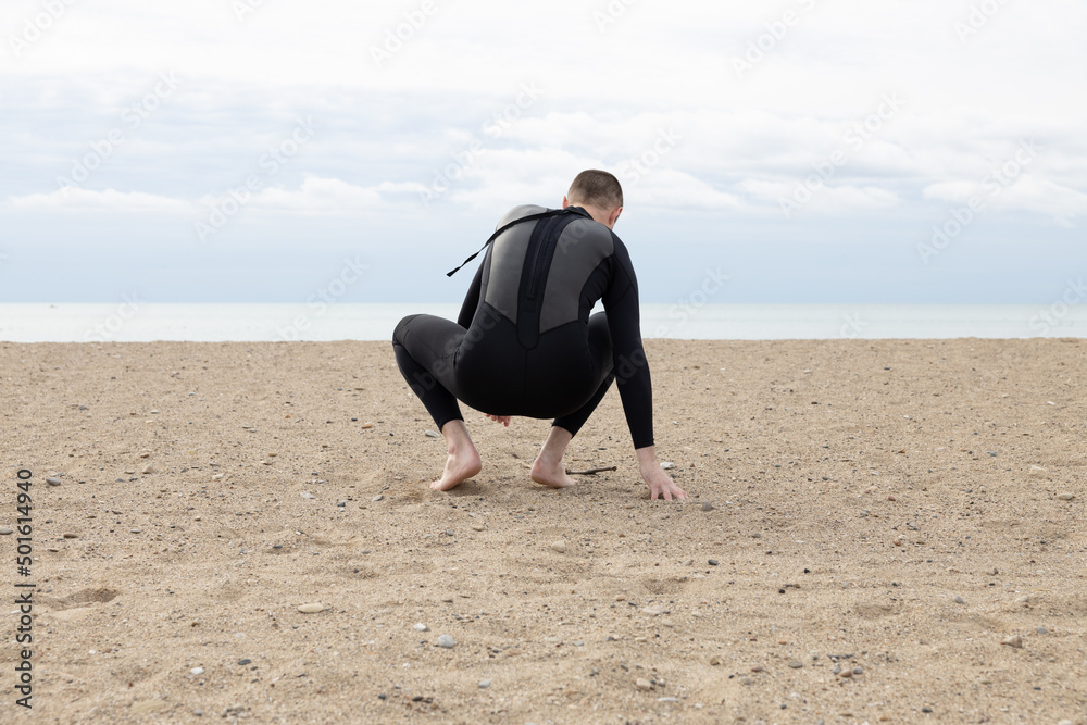 Man crouching alone on a beach next to the ocean looking out wearing a