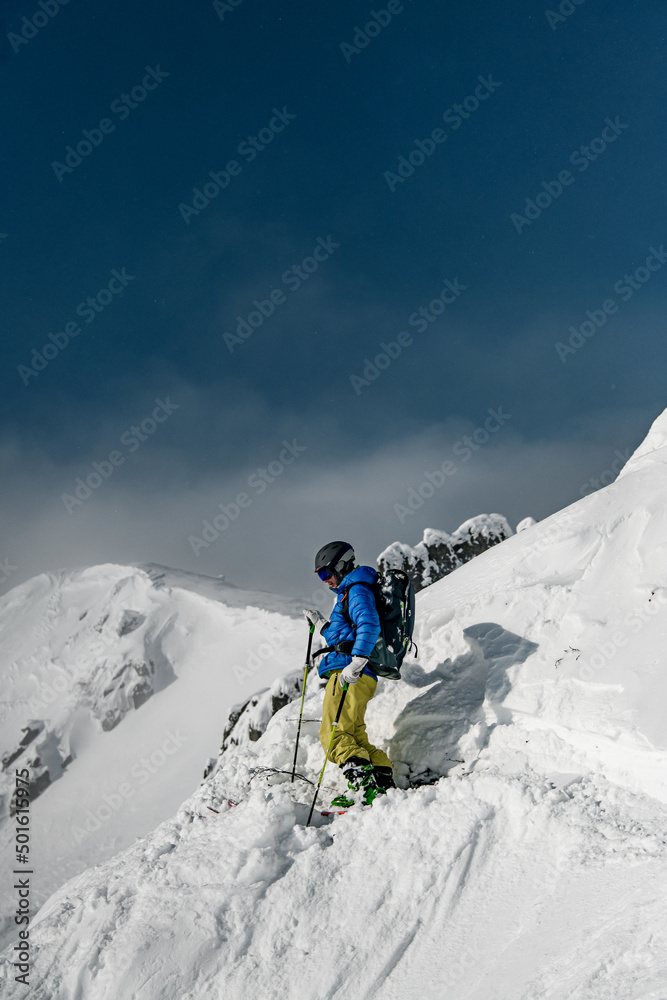 Skier man in goggles, helmet and blue and yellow ski suit doing ski touring in winter mountai
