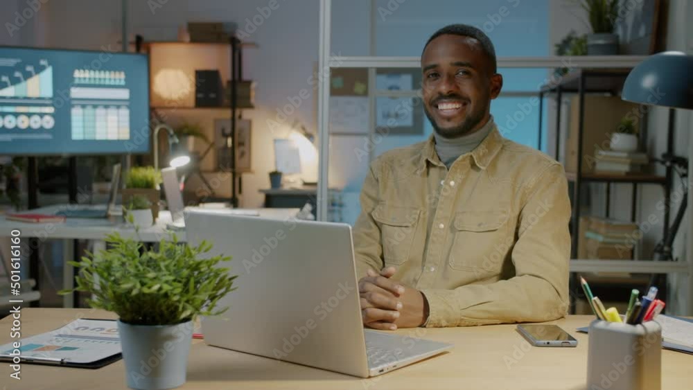 Slow motion portrait of cheerful African American businessman sitting at computer desk in office at night smiling and looking at camera Slow motion portrait of cheerful African American businessman sitting at computer desk in office at night smiling and looking at camera