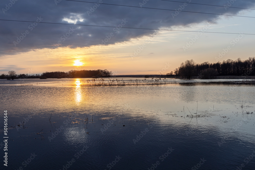 Spring flood. Sunset over the river, dramatic sky over the horizon. The sun is reflected in the water. Bright spring landscape with a river. Flooding of agricultural land.