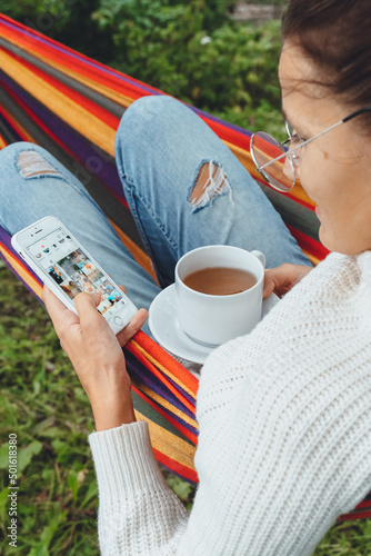 Young businesswoman use smart phone for Social media Marketing.Happy millennial check Like Follower Comment relax in hammock drink tea.Adult Influencer Outdoor browse pictures focus on device screen.