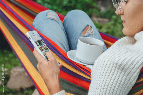 Overhead view Fall autumn millennial woman using smart phone relaxing on hammock chatting using smartphone application check social media network scrolling instagram like photos drinking tea outdoor